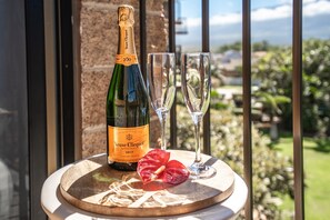 Une bouteille de champagne Veuve Clicquot Brut, deux coupes à champagne vides et une fleur rouge sur une table ronde en bois, avec en arrière-plan une vue sur la verdure et des bâtiments.
