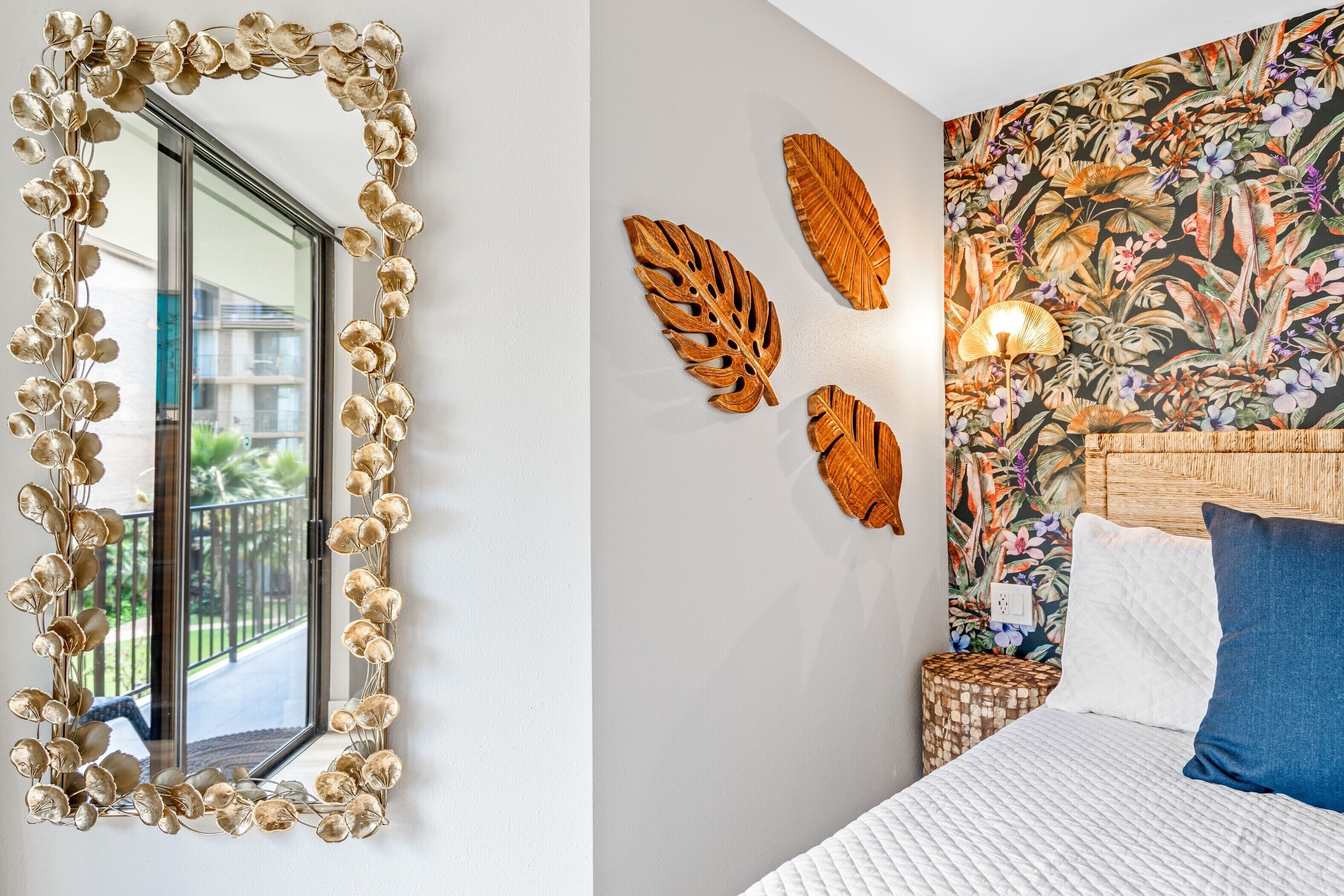 A room with a large mirror, a bed with white bedding, a floral wall tapestry, and a small side table with a lamp.