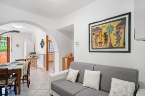 Ein Wohnzimmer mit grauem Sofa, einem Esstisch mit Stühlen, einer Küchenzeile und einem gerahmten Kunstwerk an der Wand.