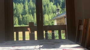 A room with a bed, a wooden table, and a view of a forest and mountains through large windows.