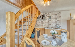 A wooden staircase leading to a loft bedroom, a kitchen with brick backsplash, and a dining area with a table set for two.