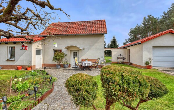 A two-story house with a red-tiled roof, a front garden with potted plants, a bench, and a small table.