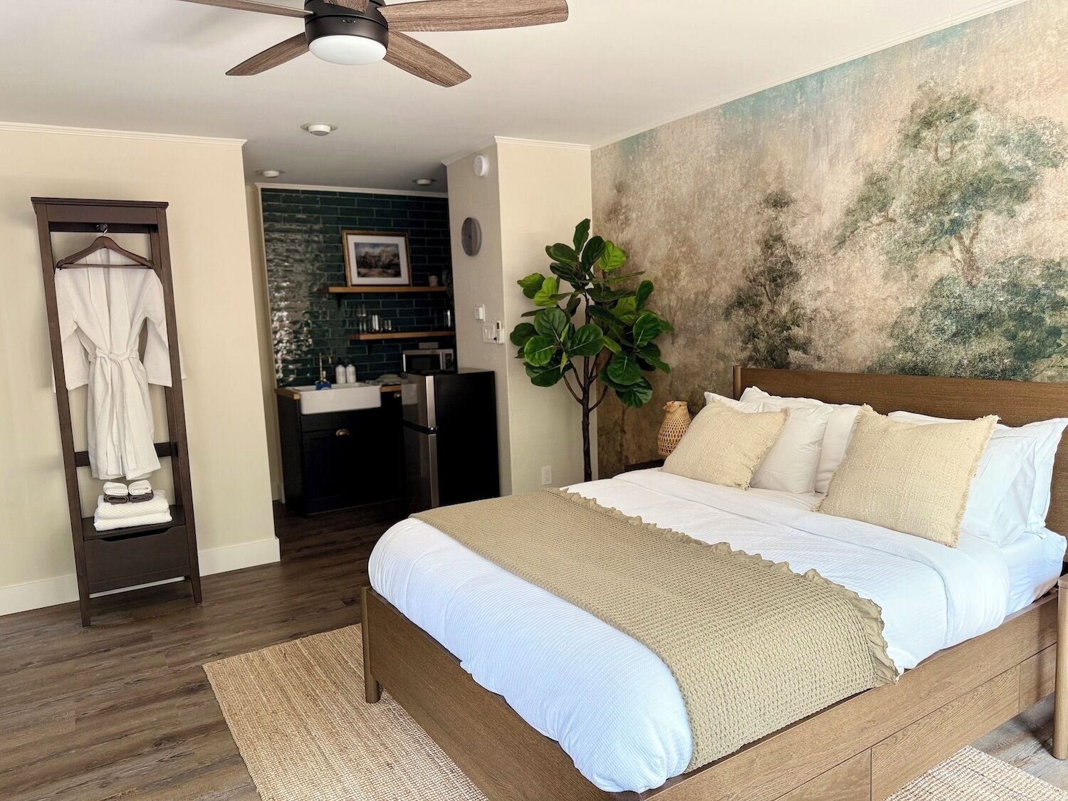 A bedroom with a wooden bed, a ceiling fan, a large plant, and a view of the bathroom.