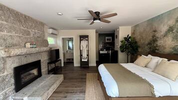 A bedroom with a stone fireplace, a bed with white bedding, a ceiling fan, and a view into the bathroom.