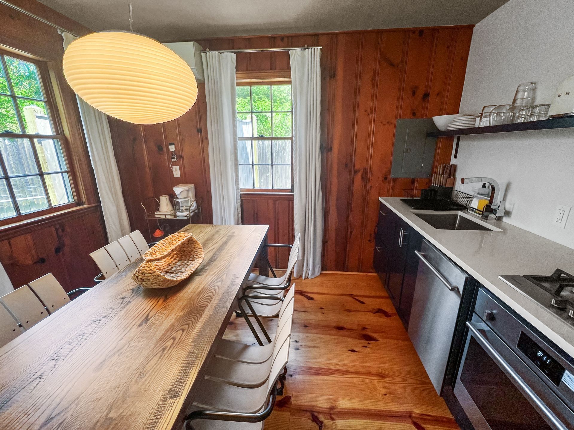 A compact kitchen with wooden cabinetry, a dining table with chairs, a hanging light fixture, and a view of the outdoors through a window.