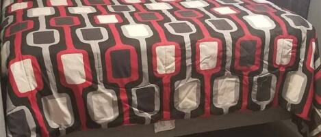 Una cama con una colcha de estampado geométrico, una mesita de noche, una lámpara y una ventana con cortinas.