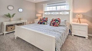 A bedroom with a white bed, two nightstands, a ceiling fan, a window with white curtains, and a small desk with a lamp.