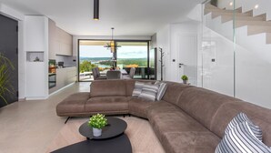 Una sala de estar moderna con un sofá seccional, un comedor con mesa y sillas, una cocina con armarios blancos, y un amplio ventanal corredero que da a un balcón con vistas al paisaje.