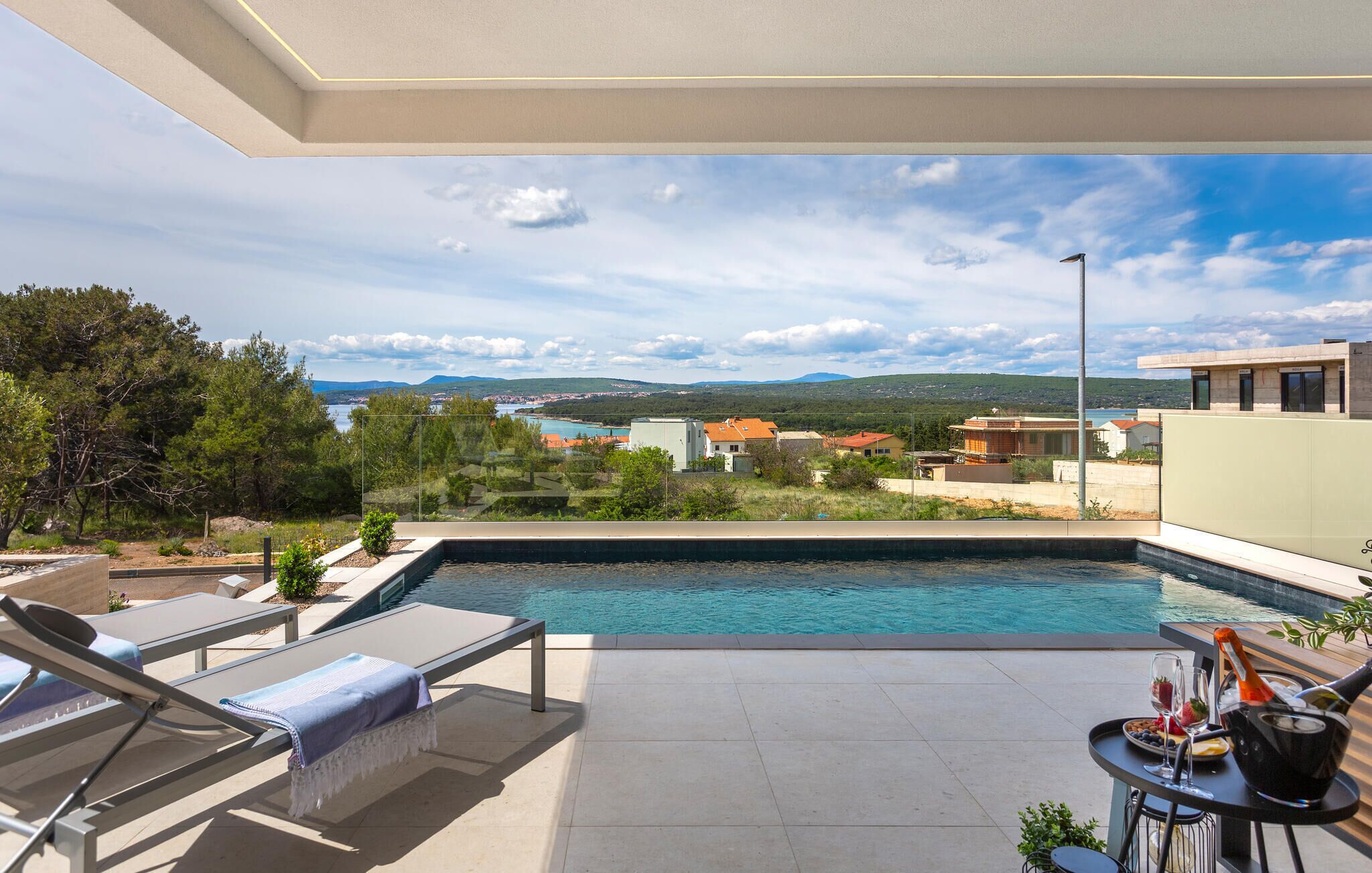 A poolside area with lounge chairs and a table with drinks, overlooking a scenic landscape with buildings and a body of water.