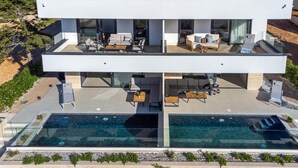 Una casa moderna de dos pisos con un amplio espacio para piscina, cerrado con vidrio, y mobiliario de exterior.