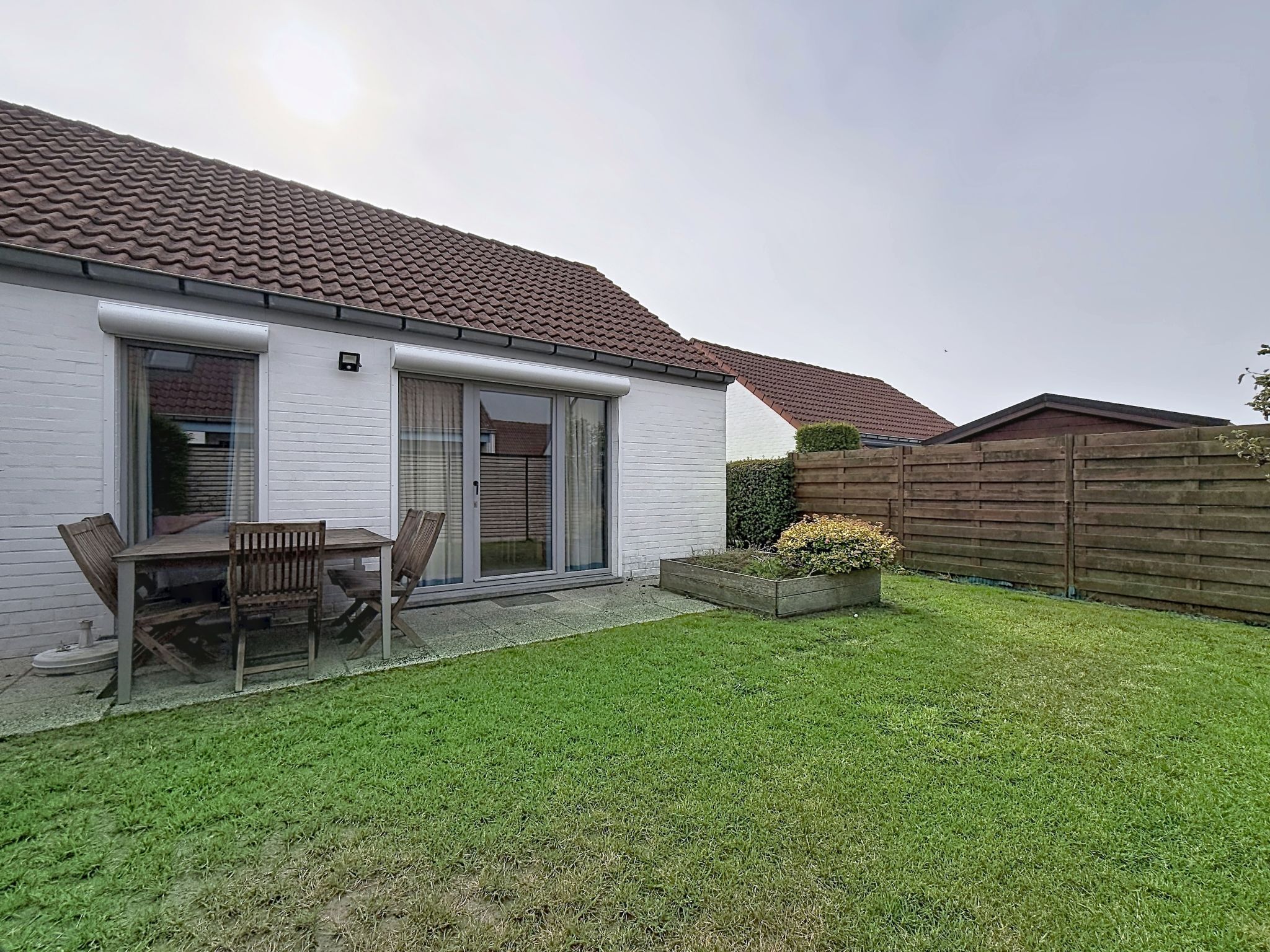 A residential house with a tiled roof, a patio with a table and chairs, a wooden fence, and a well-maintained lawn.