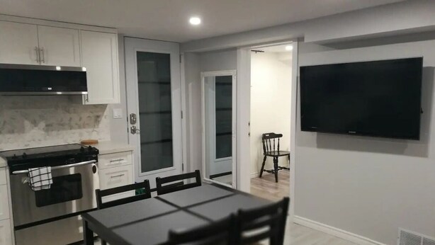 A modern kitchen with white cabinets, a black countertop, and a built-in oven. There is a dining table with chairs and a flat-screen TV mounted on the wall. - Beautiful 2-bedroom Guest Home in Guelph (Guelph)
