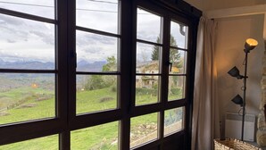 Une pièce dotée de grandes fenêtres donnant sur un paysage verdoyant et des montagnes. On y trouve une lampe et un panier posés sur le sol.