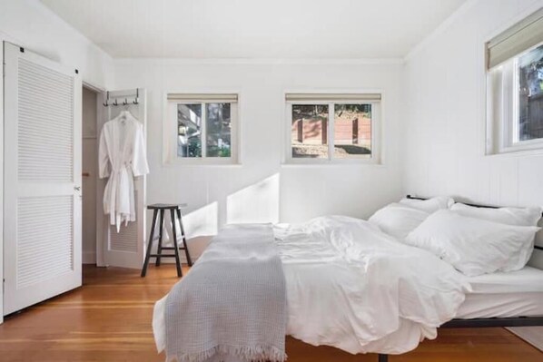 A bedroom with a bed, white walls, two windows, and a wooden floor.