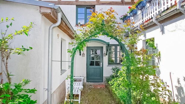 A narrow alleyway with a green archway leading to a door, surrounded by plants and a white bench. - Ferienwohnung Waren (Müritz) (Waren (Müritz))