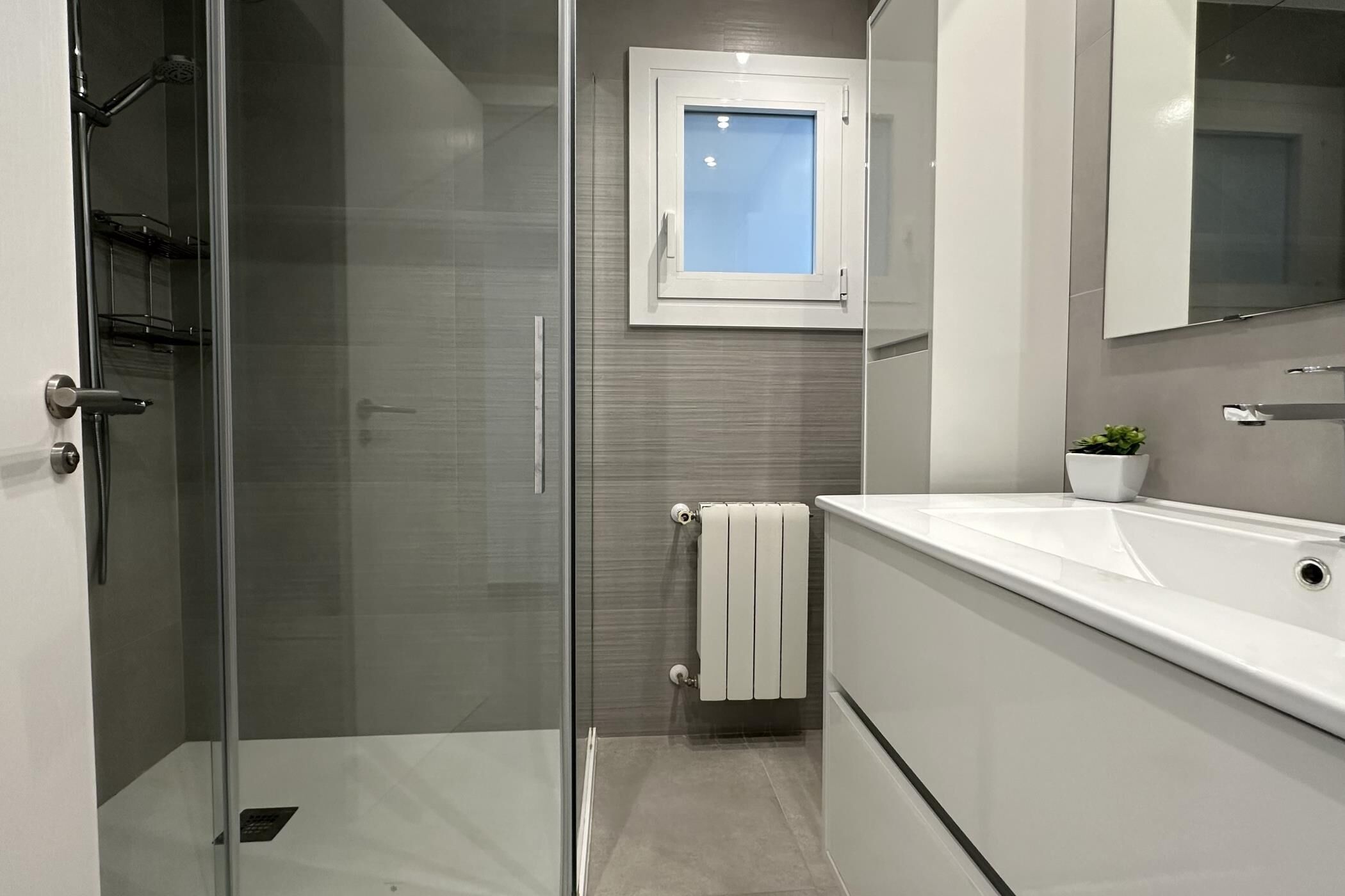 Een moderne badkamer met een douchecabine van glas, een wit wastafelmeubel en een klein raam boven de spiegel.