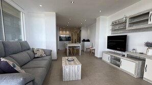 Un salon moderne avec un canapé gris, une table basse et un téléviseur posé sur un meuble en bois. On aperçoit une cuisine à l’arrière-plan, avec un coin repas.