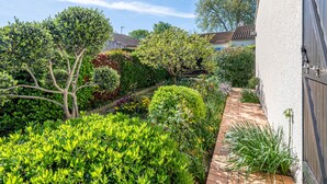 Un jardin bien entretenu, agrémenté de diverses plantes, avec un chemin en briques menant à un bâtiment blanc doté d’une porte en bois foncé.