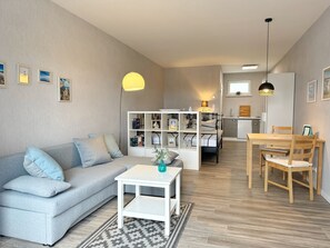 Una sala moderna con un sofá gris, una mesa de centro blanca y una estantería. Hay una zona de comedor con una mesa y sillas de madera.