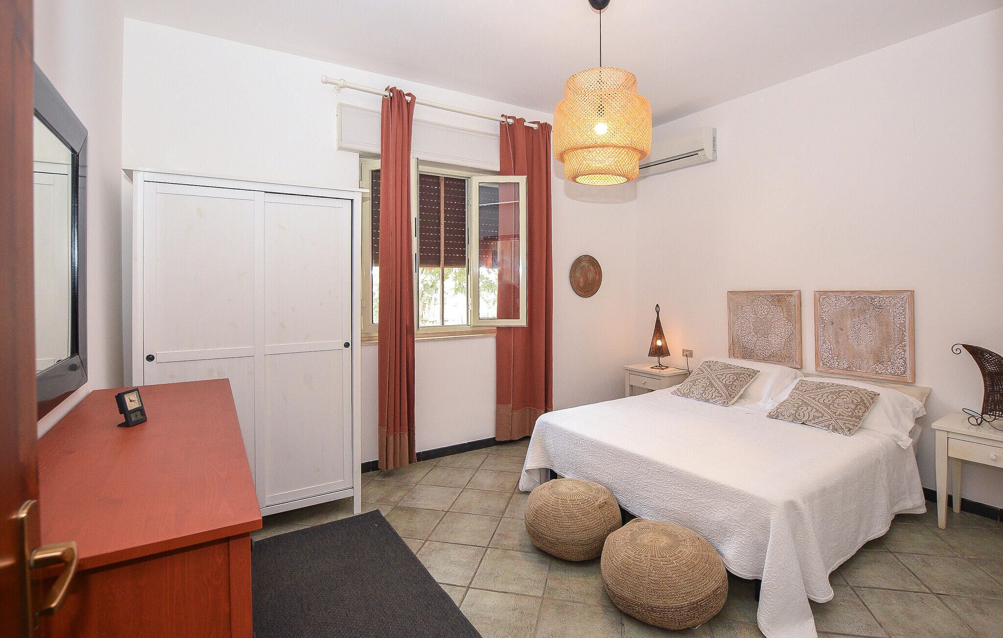 A bedroom with a bed, bedside tables, a mirror, a hanging light fixture, and a window with curtains.