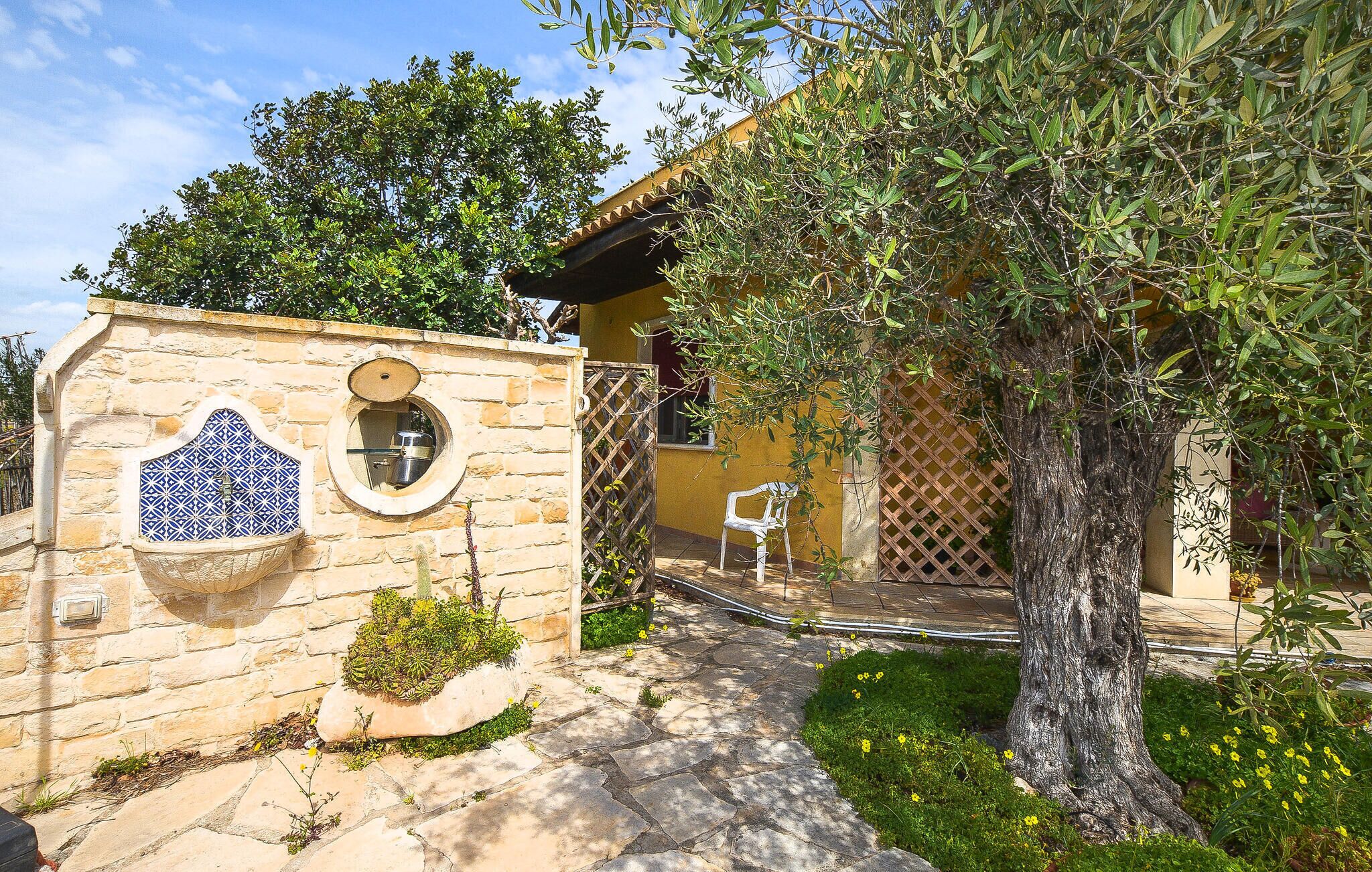 A stone wall with a circular window and a blue tile design, a tree-lined pathway leading to a building, and a white chair in the garden.