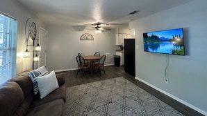A living room with a sofa, a dining table with chairs, a ceiling fan, a mounted TV, and a patterned rug.