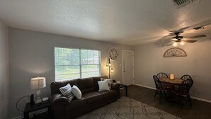 A living room with a brown sofa, a patterned rug, a dining table with chairs, a ceiling fan, and a clock on the wall.