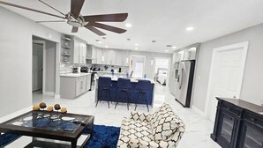 Una cocina moderna con armarios blancos, una isla central con taburetes de bar, un ventilador de techo y un comedor con mesa de vidrio y alfombra azul.