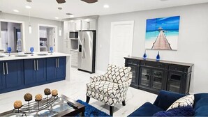 Una cocina moderna con electrodomésticos de acero inoxidable, una zona de comedor con una mesa de vidrio, y una sala de estar con un sillón estampado y un sofá azul.