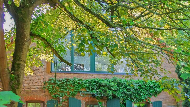 A brick building with green shutters and a tree in the foreground. - Ferienwohnung Rheinsberg (Rheinsberg OT Linow)