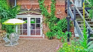 A brick building with a glass door entrance labeled "Lounge," surrounded by greenery and a gravel patio. - Ferienwohnung Rheinsberg (Rheinsberg OT Linow)