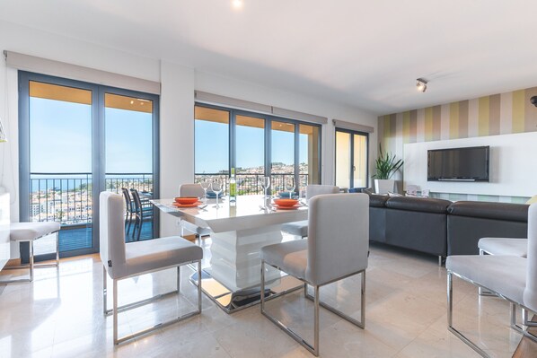 A modern kitchen with a dining area, large windows, and a flat-screen TV.