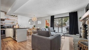 Un salon moderne avec un canapé gris, un coin repas avec une table et des chaises, une cuisine avec des meubles blancs et une grande fenêtre donnant sur des arbres.