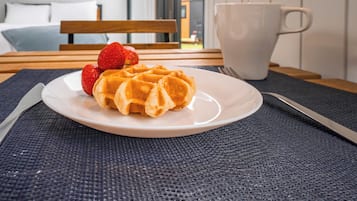Lautasella on vohveleita ja mansikoita, pöydällä valkoinen kuppi ja taustalla näkyy sänky.