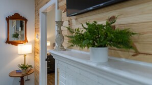 Une pièce avec une cheminée blanche, un téléviseur fixé au mur, un miroir et une petite table sur laquelle se trouvent une lampe et une plante.