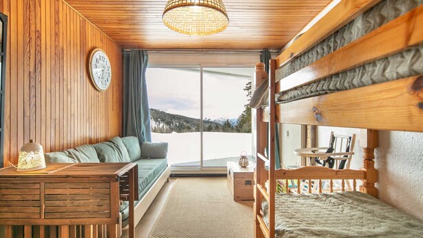 Ein gemütliches Blockhauszimmer mit Holzausstattung, einer Couch, einem Etagenbett, einem Schreibtisch und einem großen Fenster mit Blick auf die Berge.