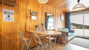 Ein gemütliches Zimmer mit Holzwänden, einem runden Esstisch, klappbaren Stühlen, einer Couch und einem großen Fenster mit Blick, sowie einer Wanduhr.