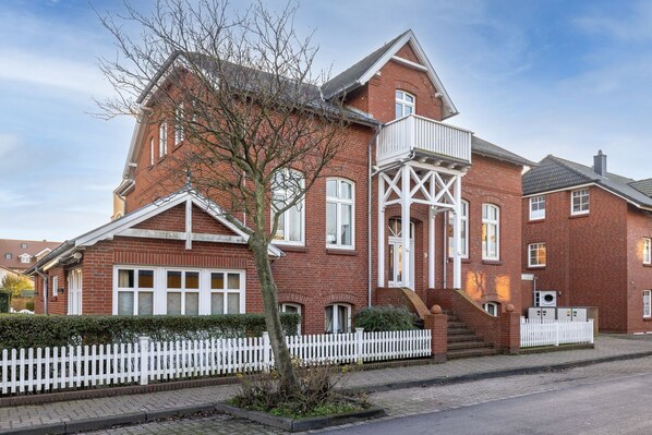 A red brick building with a white picket fence, a balcony, and a staircase leading to the entrance.
