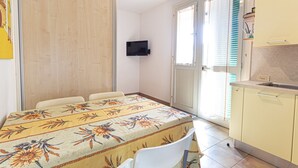 A small, bright room with a dining table, chairs, a TV mounted on the wall, and a kitchen area with cabinets and a sink. - Two-room apartment Lina, ground floor with fenced outdoor space near the center of Marina di Campo (Campo nell'Elba)