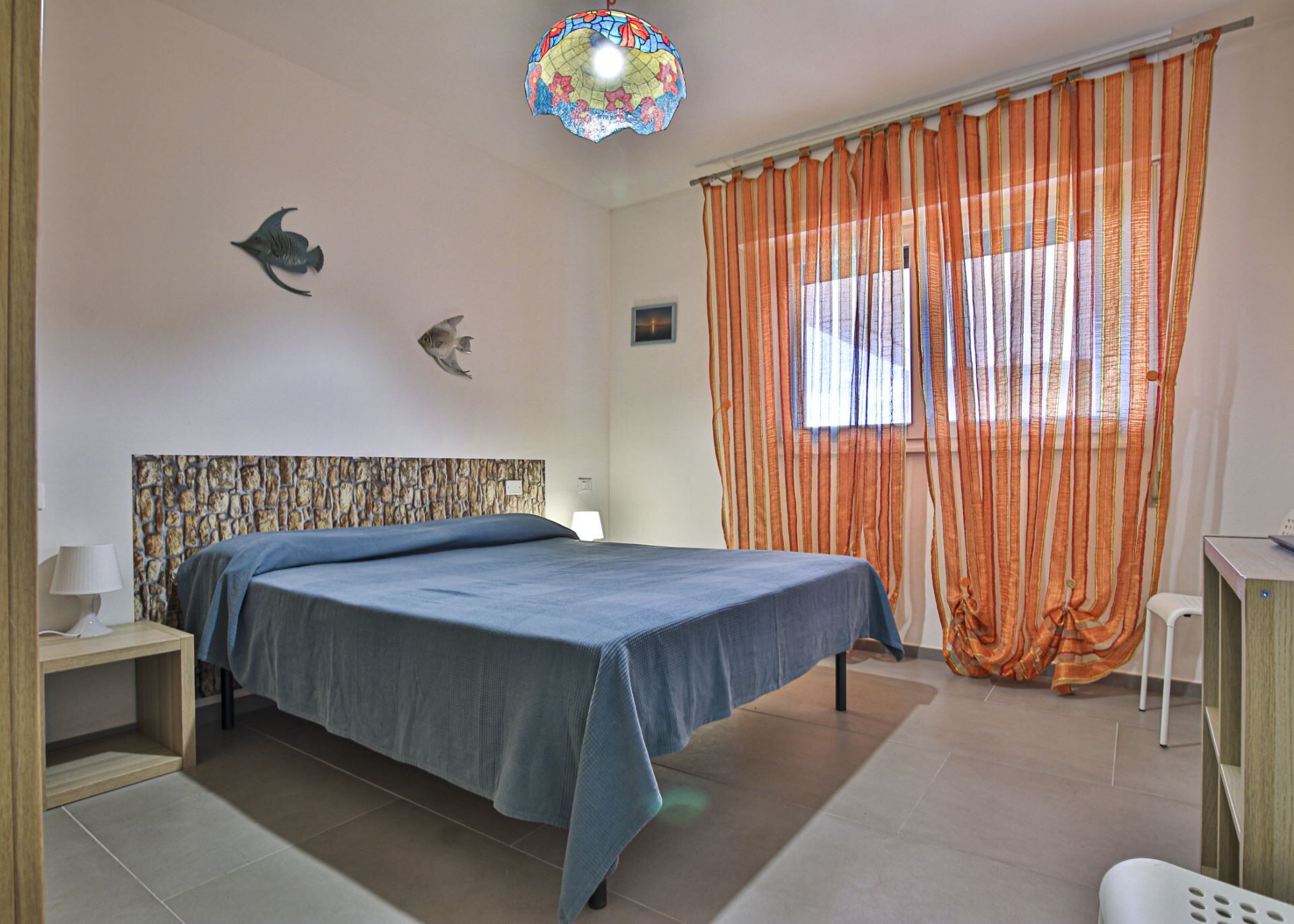 Una camera da letto con un letto, un comodino, una lampada, un'opera d'arte a tema pesci, una lampada a soffitto in vetro colorato e tende arancioni.