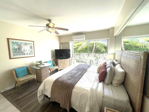 A hotel room with a bed, a desk, a chair, a ceiling fan, and a large window with a view of greenery.