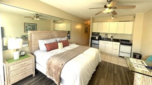 Un dormitorio con cama, mesitas de noche, espejo, ventilador de techo y una kitchenette con estufa y microondas.
