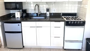 Cocina compacta con armarios blancos, encimera negra, microondas, tostadora, lavabo, cafetera, estufa a gas y refrigerador.