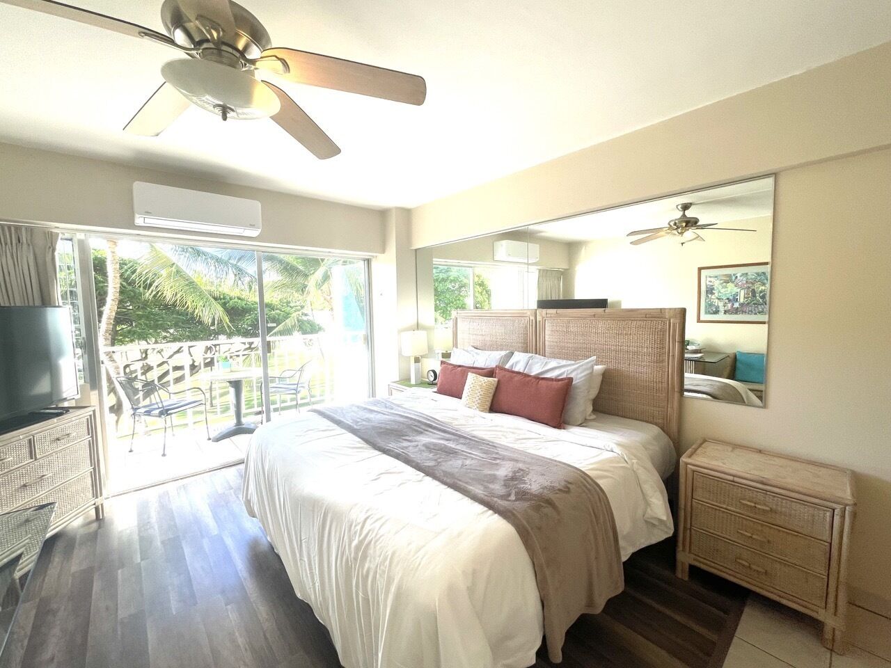 A bedroom with a bed, a nightstand, a TV, a ceiling fan, and a balcony with a view of greenery.