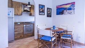 A compact kitchen with wooden cabinets, a gas stove, and a refrigerator. There is a dining area with a round table and chairs, and a painting of a coastal scene on the wall.