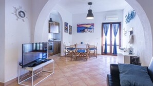 A living room with a TV, a sofa, a dining area with chairs, and a kitchen in the background.