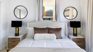 Cama bem arrumada com roupa de cama branca, dois espelhos redondos nas mesas de cabeceira e uma janela com cortinas.