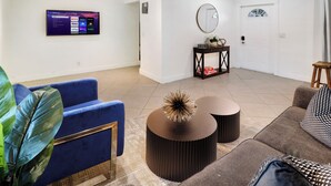 Uma sala de estar moderna com televisão de tela plana, sofás azuis e marrons, uma mesa de centro redonda e uma pequena mesa lateral com objetos decorativos.
