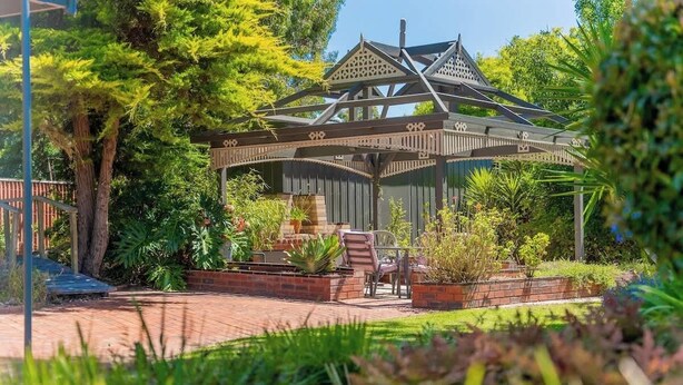 Un belvédère doté d’un toit soutenu par des poutres en bois, entouré d’un jardin où poussent différentes espèces de plantes et bordé d’un chemin en briques.