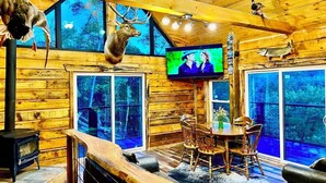 A log cabin interior with a fireplace, wooden dining table, chairs, and a television mounted on the wall.